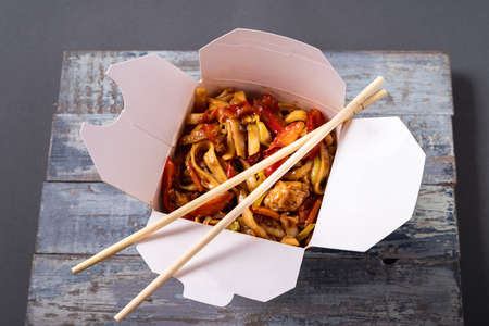 Noodles with chicken and vegetables in a box on wooden background and chopsticks, top view.の写真素材