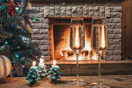 Christmas eve. Two glasses of champagne wine before Christmas tree decorated toys and christmas lights near cozy fireplace, in country cottage.の写真素材
