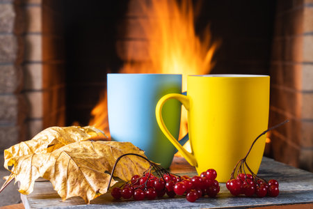 Two mugs of tea or coffee, autumn leaves and viburnum berries near a cozy fireplace, in a country house, autumn or winter holidays, horizontal.の写真素材