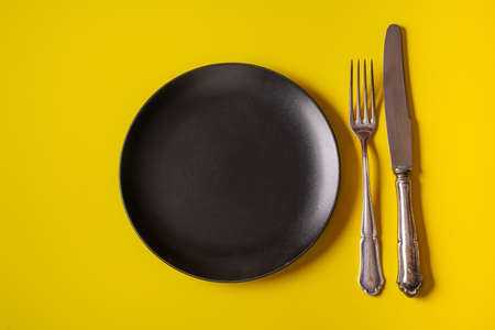 Minimalism style. Empty black plate and a cutlery set on yellow paper background.の写真素材