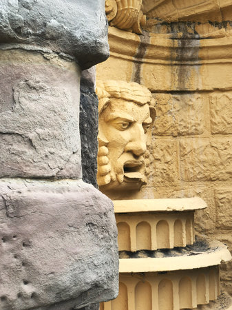 Classical stone sculpture in a historic fountain. The sculpted face, with intricate details, is set against a weathered yellow wall, partially hidden by rough stone blocks.の写真素材