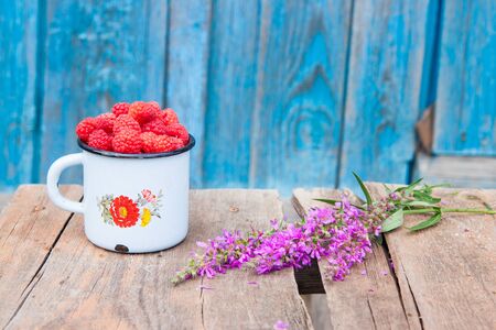 Old enamel cup with handful of fresh rasperries and a bunch violet field flowers on rustic wooden table.の写真素材
