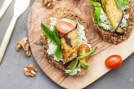 Bruschetta with grilled eggplant, cherry tomatoes, cheese dip, fresh basil and aromatic herbs on a wooden board. Healthy mediterranean cuisine.の写真素材