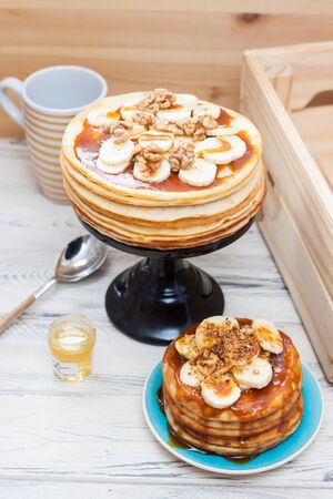 Stack of fluffy pancakes with maple syrup, banans, jam and nuts. Rustic wooden background. Cup of...