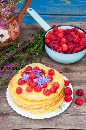 Crepes with fresh raspberry, purple field flowers and bowl with berries on rustic wooden table in front of wooden blue wallの写真素材