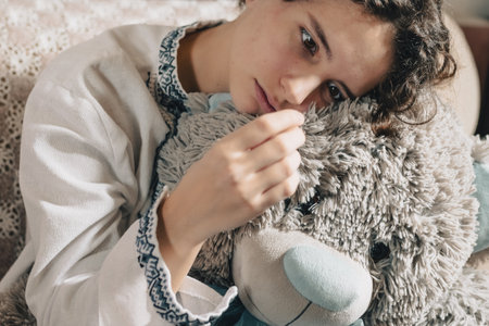 teenage girl with brown curly hair, in white ethnic embroidered shirt, with feeling of emptiness and hopelessness, in state of psychological stress or panic attack, sits hugging toy bear. sad teenagerの写真素材