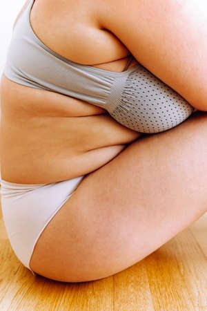 Unrecognizable curvy woman in white panties and frameless bodysuit sits on wooden parquet floor, demonstrates concept of positive body and self-acceptanceの写真素材