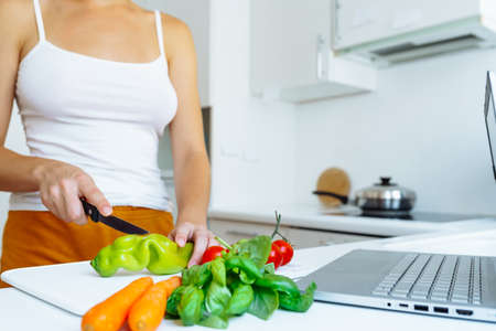 young woman with slim figure, in tank top and shorts, prepares fresh vegetable salad, according to...