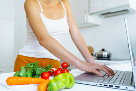 young woman with slim figure, in tank top and shorts, prepares fresh vegetable salad, according to online recipe, in home kitchen. healthy food for breakfastの写真素材