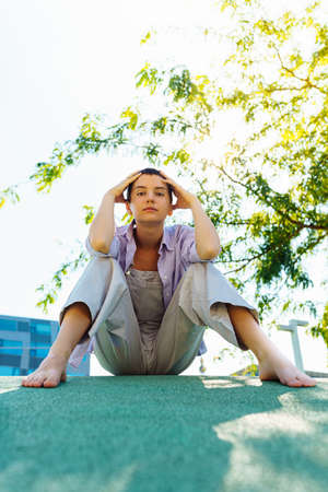 Portrait of teenage girl with short hairstyle, in unisex clothing, sitting barefoot on blurred background of urban architecture, clasping head in hands, on artificial turf. large vの写真素材