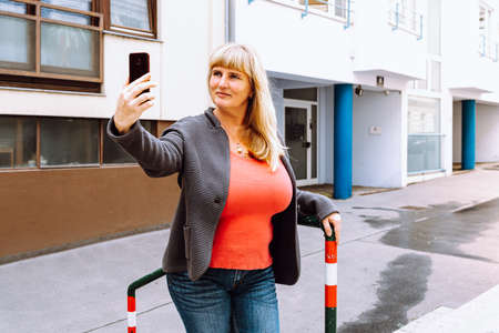 Mature happy middle aged woman, 40s, with long blond hair, smiling, wearing jeans and knitted jacket, talking on smartphone, on video call, taking selfieの写真素材