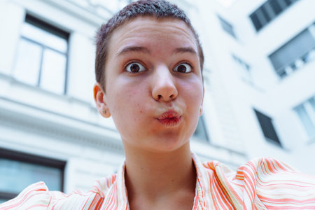 Selfie portrait of teenage girl with short hairstyle, teenage acne, kiss-shaped lips, looking at camera on blurred background of building with windows, school or universityの写真素材