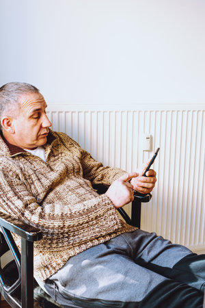 middle-aged man with big fat belly, in warm knitted sweater, sits on chair near heating radiator, rummages through mobile Internet. concept of early aging and depression in the middleの写真素材