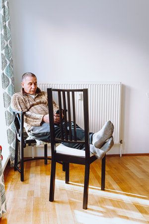 middle-aged man with large fat belly sits on chair near heating radiator, with feet on another chair, against background of window and nearby sports weights.の写真素材