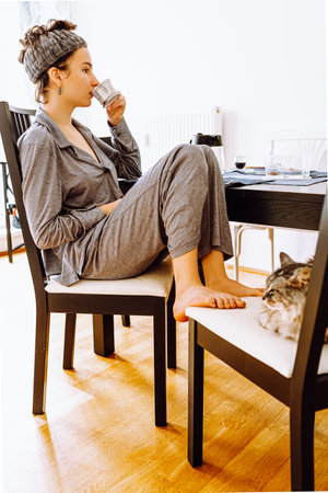 teenage girl, with hairband on head, in gray pajamas, sits on chair, bare legs tucked under her, drinks coffee in sunny morning dining room, domestic cat sits on chair next to her.の写真素材