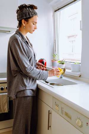 girl teenager in pajamas, barefoot on parquet floor, in kitchen, washes dirty burnt frying pan in...