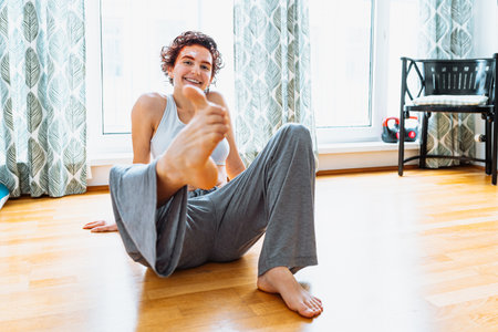 positive yoga at home. teenager girl with red-dyed hair and eyebrows, cream mask on face, in pajamas and top, shows ok symbol with big toe, sitting on parquet floor at home.の写真素材
