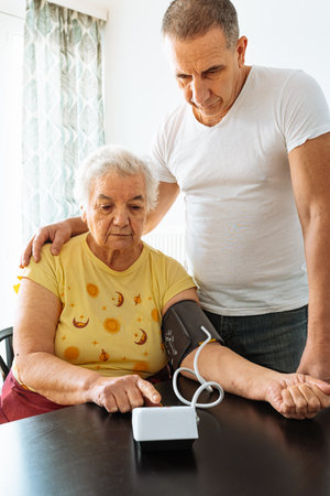 elderly woman with hypertension measures blood pressure home. middle-aged man, son stands nearby and carefully hugs mother.の写真素材