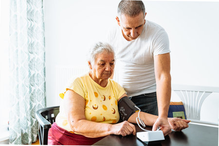 elderly woman with hypertension measures blood pressure home. middle-aged man, son stands nearby and carefully hugs mother.の写真素材