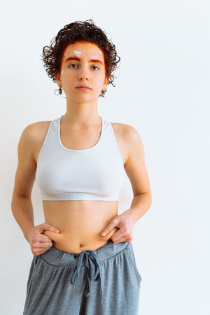 young teenage girl, with bright red hair dye on hair and eyebrows, cream mask on face, in gray top and pajama pants, demonstrates saggy skin on stomach, overweight and fat foldsの写真素材