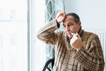 Middle-aged man in warm sweater, sits on an armchair, measures temperature, suffers from headache....