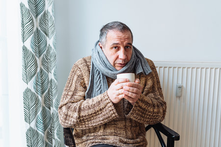 middle-aged man with cold, virus or flu, with runny nose, in warm sweater and woolen scarf, drinks warm drink from cup, an unhappy expression on face.の写真素材