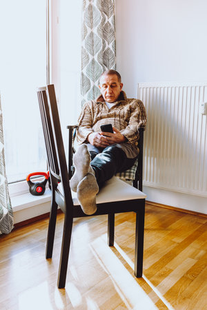 middle-aged man with large fat belly sits on chair near heating radiator, with feet on another chair, against background of window and nearby sports weights. concept of early agingの写真素材