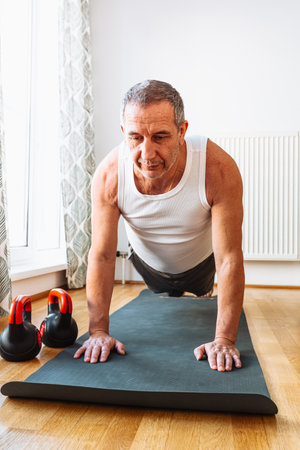middle-aged man, muscular build, in tank top and sports shorts, stands in plank position, does strength exercises, with weights on sports mat, at home against blurred background ofの写真素材