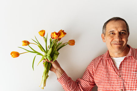 Portrait of positive smiling middle-aged man, in plaid shirt, with bouquet of tulips in hand against light wall. Man with gift bouquet positive emotionの写真素材