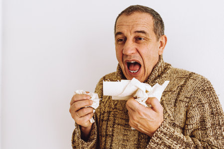 Middle-aged man in warm sweater, blows nose into disposable paper napkin, sneezes and coughs, is sick at home. Man suffering from cold sneezes and blows noseの写真素材