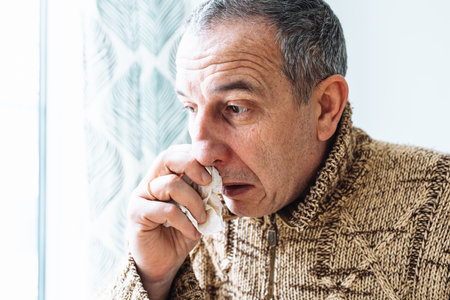 Middle-aged man in warm sweater, blows nose into disposable paper napkin, sneezes and coughs, is sick at home. Man suffering from cold sneezes and blows noseの写真素材