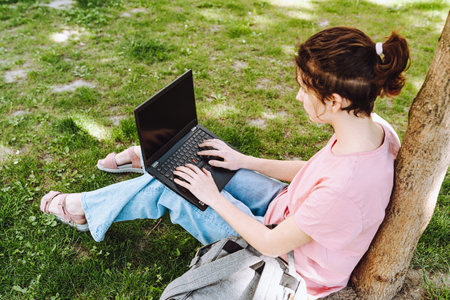 back view of young woman using laptop while sitting on grass in parkの写真素材