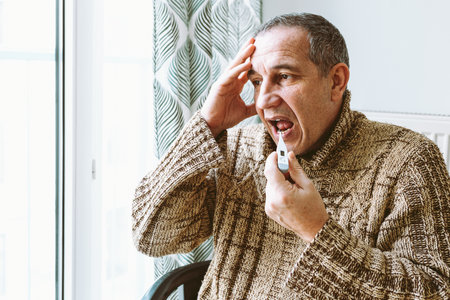 middle-aged man with headache and fever measures temperature with an electronic thermometer. Sits on chair near window in warm knitted sweaterの写真素材