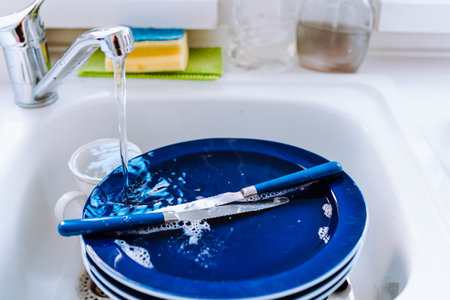Washing dishes in the kitchen sink with a brush and sponge. Water flowing from tap onto dirty dishes in kitchen sink. Bright blue plates stacked after breakfastの写真素材