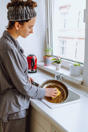 young woman, teenager, daughter, in gray pajamas, with bandage on brown hair, washes dishes in sink in small modern kitchenの写真素材
