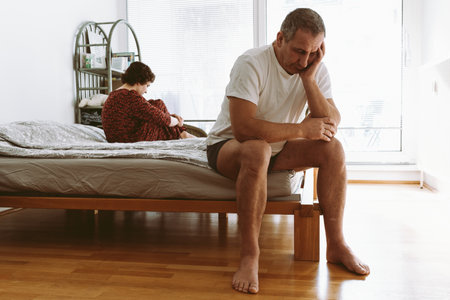 middle-aged man sits on edge of bed in a deplorable state, propping head in hands, young woman sits on opposite edge. Quarrel in bedroomの写真素材