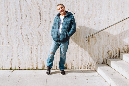 Full length portrait positive middle-aged man, in warm winter jacket, jeans, sneakers, against background marble stone wall and stairs with railingの写真素材