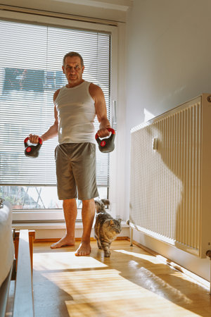 Senior man lifting dumbbells while standing by the window at homeの写真素材