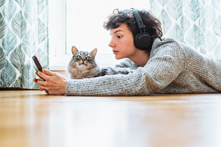 Young woman with headphones lying on the floor with cat and using smartphoneの写真素材