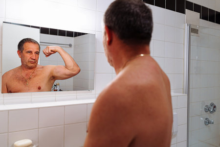 Man in bathroom looking at himself in the mirror and flexing bicepsの写真素材