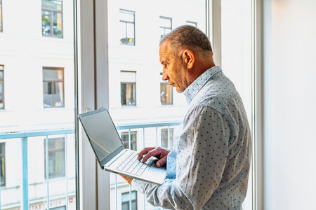 Attractive man in shirt standing near window with laptopの写真素材