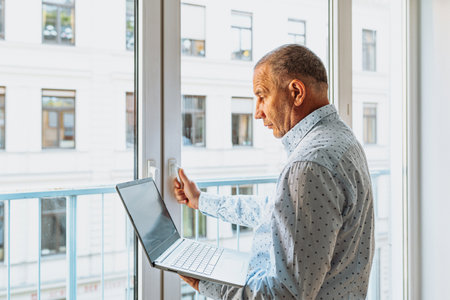 Attractive man in shirt standing near window with laptopの写真素材