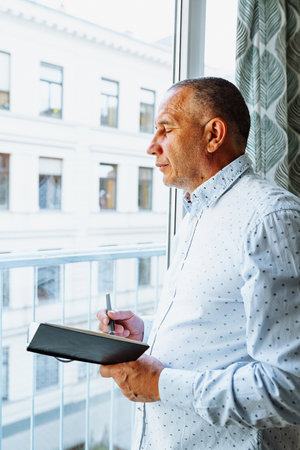Attractive man in shirt standing near window with notepadの写真素材