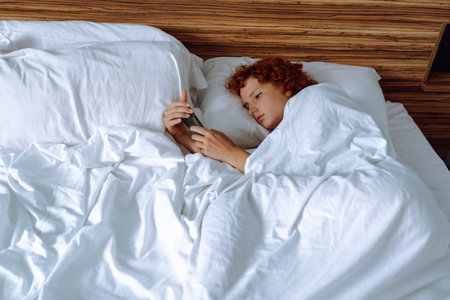 young red-haired girl in bed using gadgetの写真素材