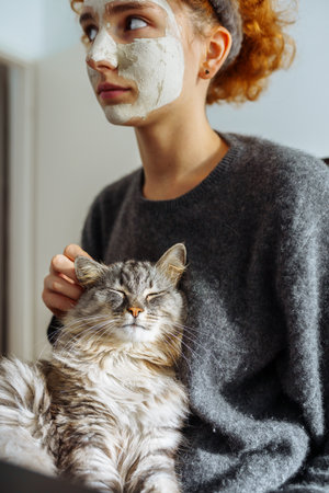 gray fluffy tabby cat sits on arms teenage girl wearing cosmetic maskの写真素材