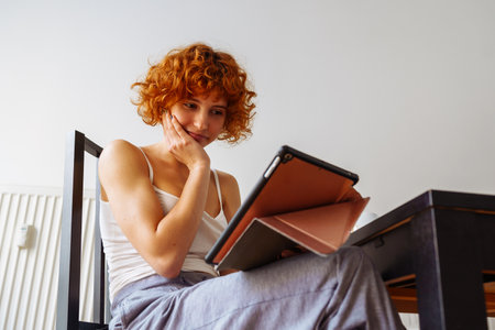 red-haired teenage girl sits in dining room at table, uses tablet, chats
