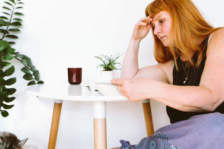 portrait middle-aged blonde woman sitting at table with book in home interiorの写真素材