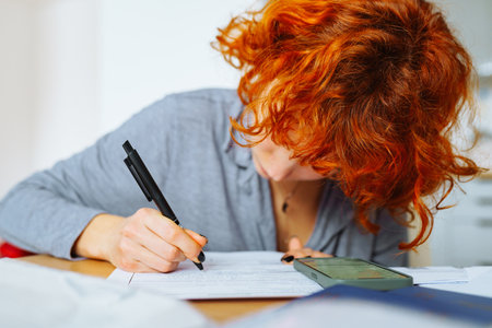 young woman, student, with red curly hair, fills out documents sitting at table at home, in...