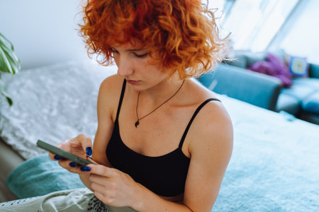 portrait of young woman with curly red hair sitting on bed in bedroom using phoneの写真素材