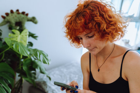 portrait of young woman with curly red hair sitting on bed in bedroom using phoneの写真素材